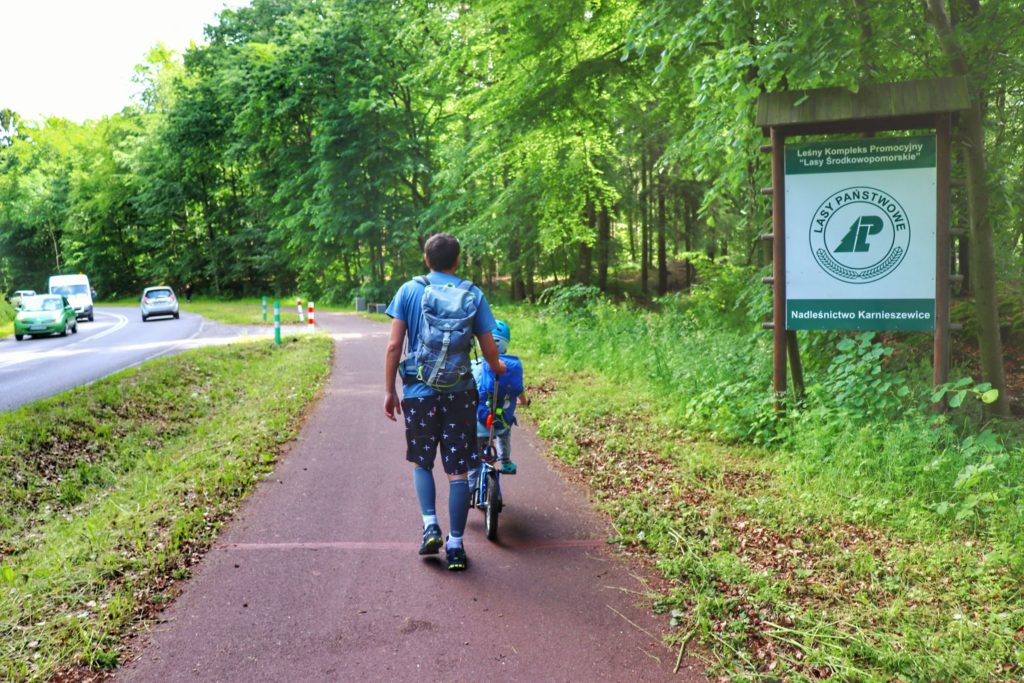 Turysta z dzieckiem jadącym na rowerze, Tablica w kolorze zielono - białym z napisem Lasy Państwowe, Leśny Kompleks Promocyjny Lasy Środkowopomorskie