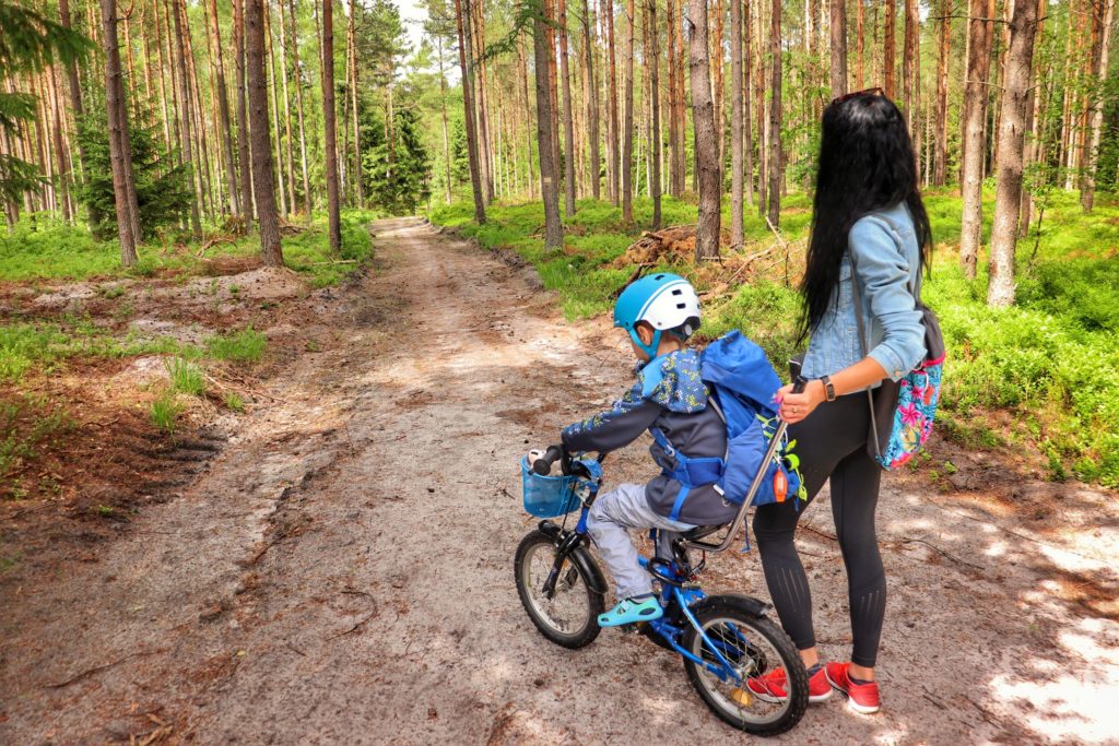 Turystka pchająca rower, na którym siedzi dziecko, szeroka droga leśna, szlak żółty w Koszalinie, słoneczny, letni dzień