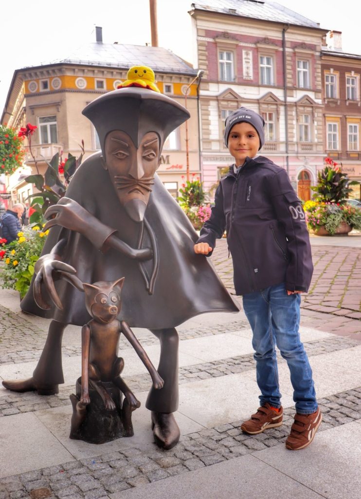 Uśmiechnięte dziecko pozujące przy pomniku Szpiega z Krainy Deszczowców -  Don Pedra de Pommidore stojąca w mieście Bielsko-Biała na ulicy 11 Listopada