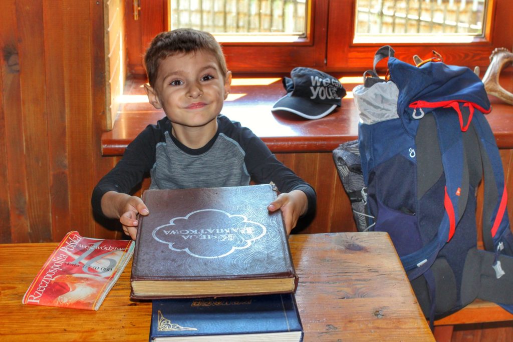 Uśmiechnięte dziecko siedzące przy drewnianym stole w schronisku na Luboniu Wielkim, trzymające w dłoniach księge gości odwiedzających schronisko