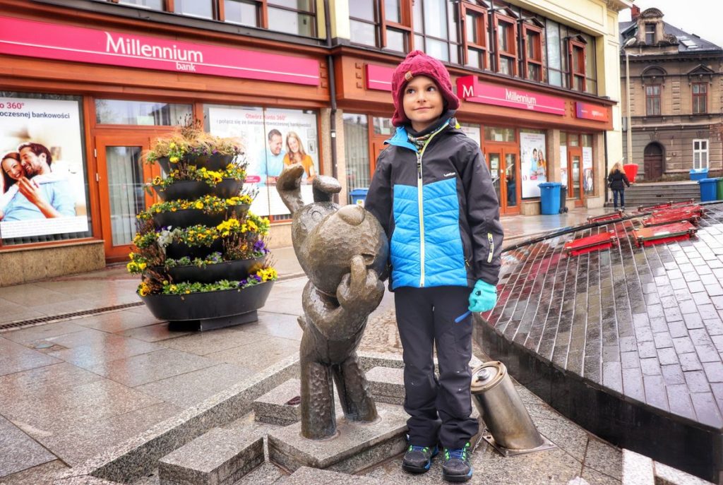 Zadowolone dziecko pozujące przy wylanej z brązu rzeźbie Reksia oraz fontannie w Bielsku-Białej przy ulicy 11-Listopada
