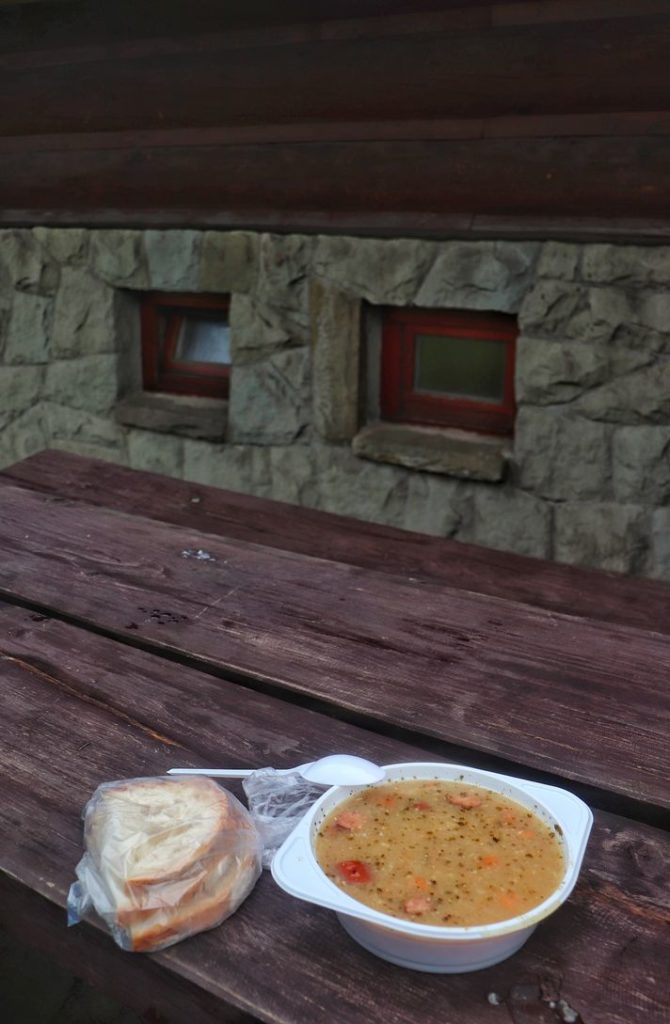 Biała, plastikowa miska z zupą - żurek, chleb zapakowany w woreczek foliowy, drewniany stół przy Bacówce na Hali Rycerzowej