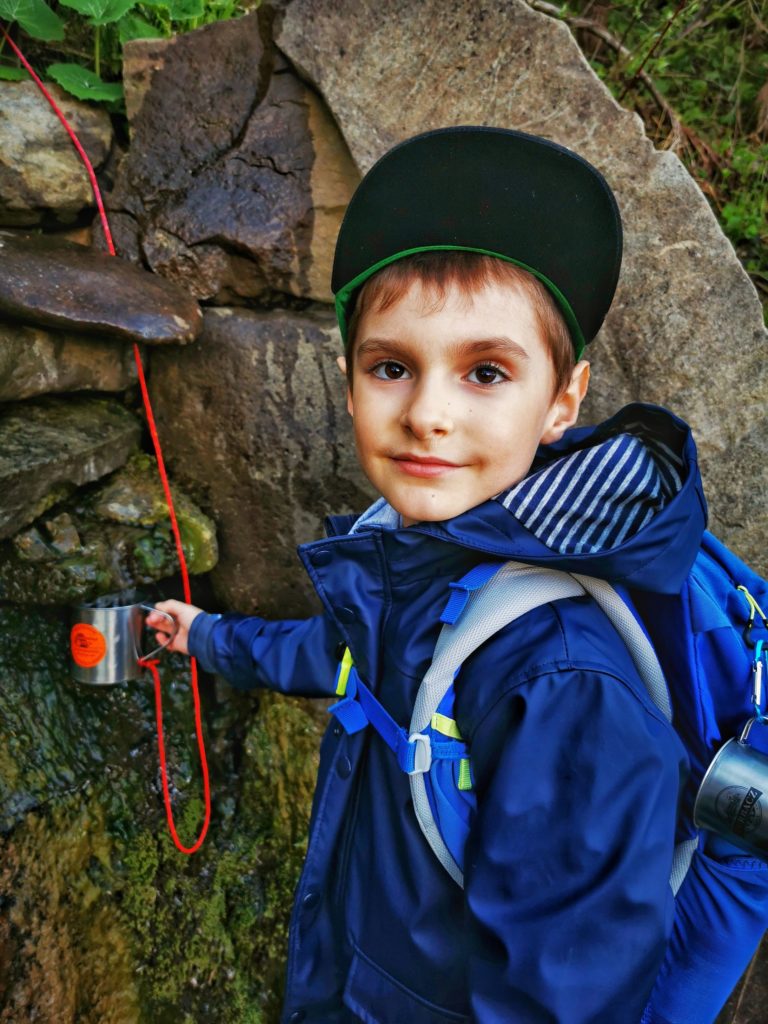 Zadowolone dziecko (chłopiec) nabierający wodę górską do metalowego kubka na czarnym szlaku na Wielką Rycerzową idącym z Soblówki