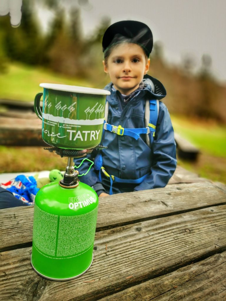 Zielona butla gazowa, na palniku postawiony jest zielony kubek z białym napisem Tatry, drewniany stół, w tle zadowolone dziecko - Hala Rycerzowa