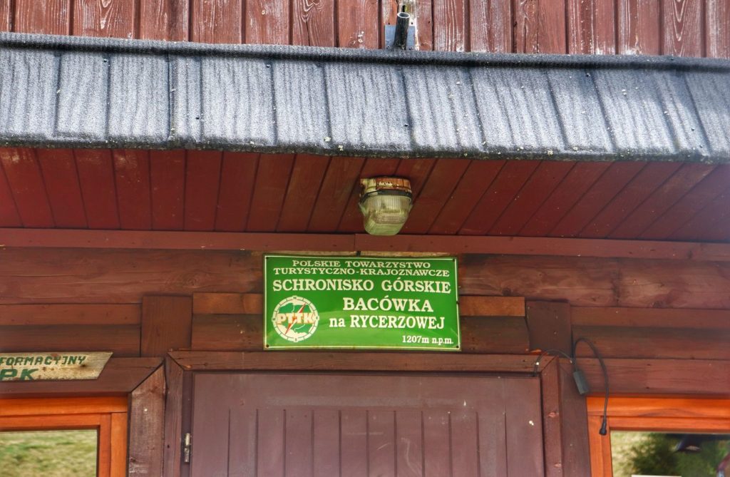 Zielona tabliczka wisząca nad drzwiami schroniska na Wielkiej Rycerzowej z napisem - SCHRONISKO GÓRSKIE BACÓWKA NA RYCERZOWEJ - 1207 M N.P.M.