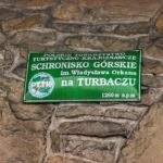Zielona tabliczka wisząca na kamiennej ścianie schroniska górskiego im. Władysława Orkana na Turbaczu 1260 m n.p.m.
