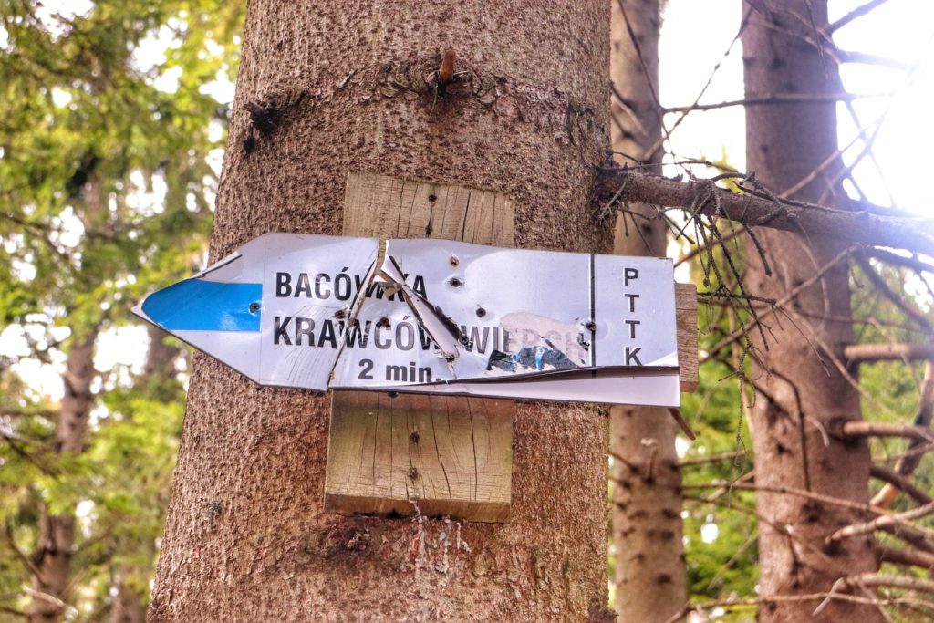 Biała tabliczka wskazująca przebieg szlaku niebieskiego do Bacówki na Krawcowym Wierchu oraz czas 2 minuty
