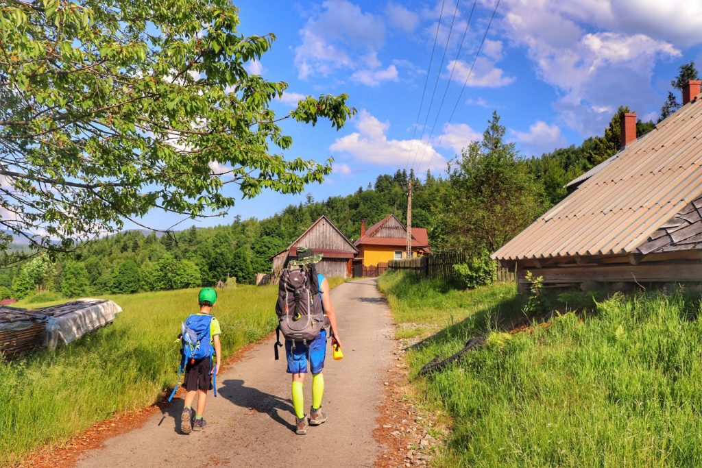 Turyści, dziecko z tatą na szerokiej drodze asfaltowej idącej między zabudowaniami w Milówce, niebieskie niebo