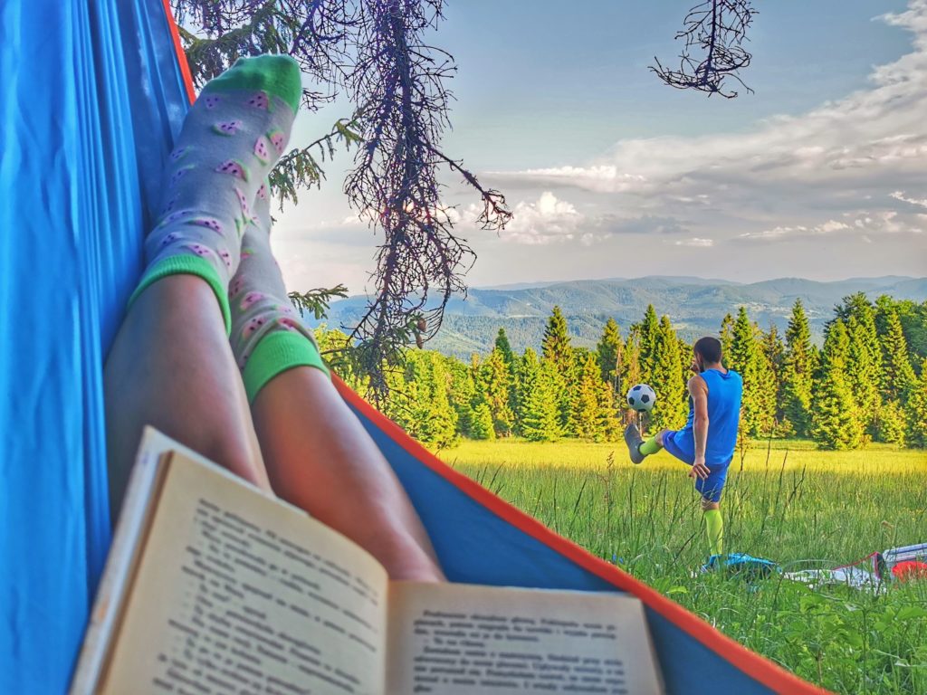 Turystka leżąca w niebieskim hamaku, czytająca książkę, w tle mężczyzna kopiący piłkę do nogi, Polana Sucha Góra w Beskidach