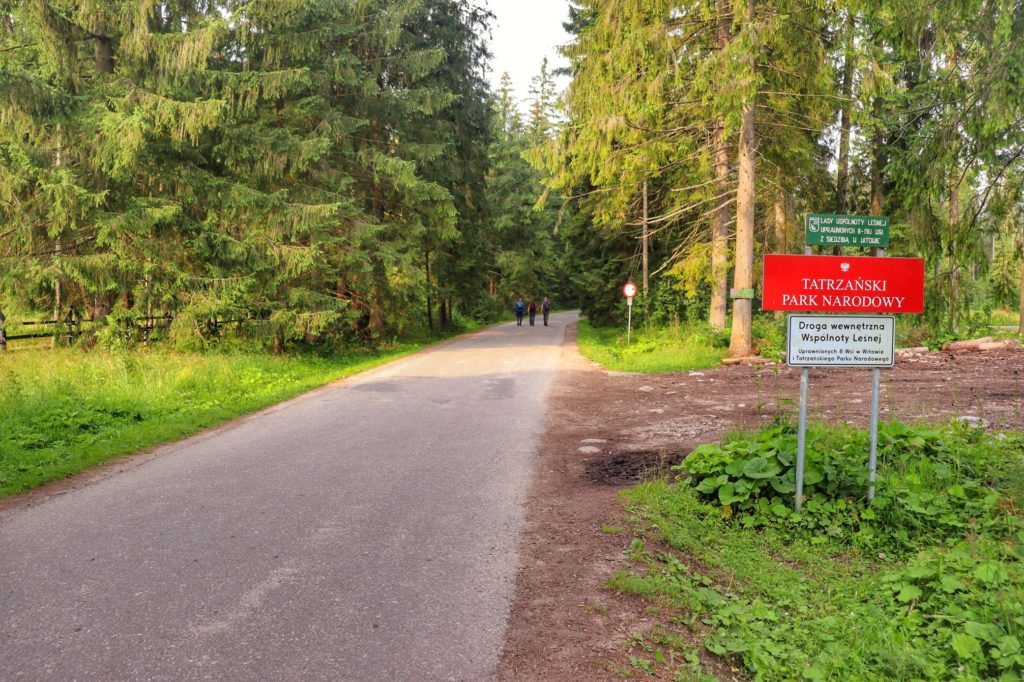 Czerwona tablica z białym napisem TATRZAŃSKI PARK NARODOWY stojąca po prawej stronie szerokiej drogi biegnącej do schroniska na Polanie Chochołowskiej w Tatrach, w oddali widoczna jest grupka turystów, lato