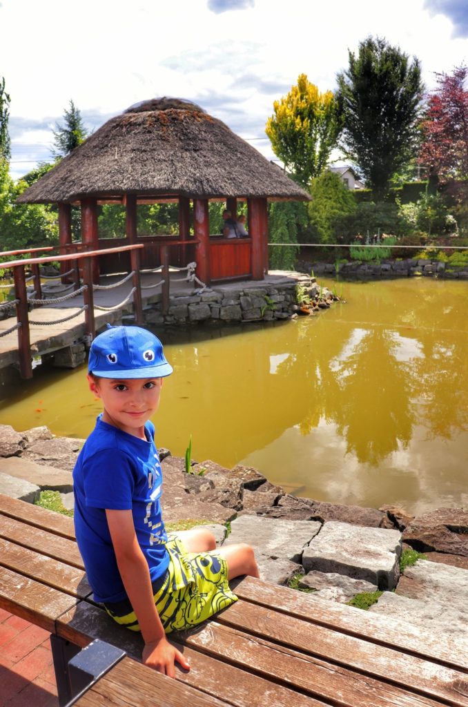 STAW Z ALTANĄ - Ogród do zwiedzania w Goczałkowicach, uśmiechnięte dzecko siedzące na drewnianej ławce przy stawie