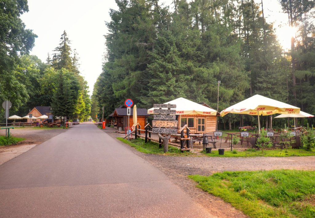 Szeroka asfaltowa droga idąca przez Dolinę Chochołowską, drewniana chatka, w której sprzedawane jest piwo, stoliki, parasolki, słoneczny poranek