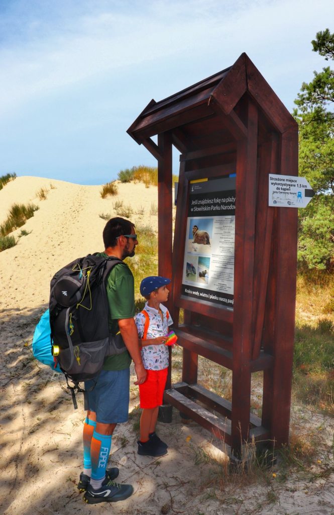 Turysta z dzieckiem, tablica informacyjna - temat FOKI znajdująca się przy wejściu na plażę w Słowińskim Parku Narodowym
