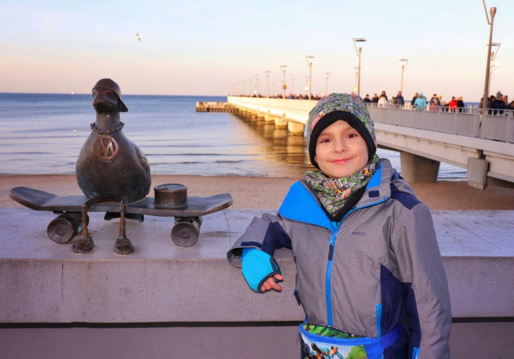 Uśmiechnięte dziecko pozujące przy figurce mewy - Marian na deskorolce w Kołobrzegu, w tle kołobrzeskie molo
