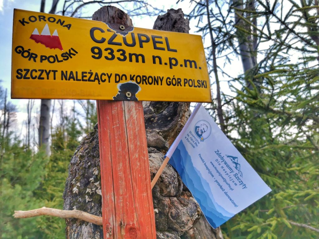 Żółta tabliczka oznaczająca najwyższy szczyt Beskidu Małego należący do Korony Gór Polski - Czupel, flaga akcji zdobywamy szczyty dla hospicjum