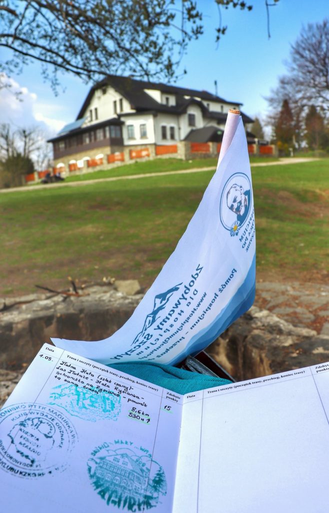 Flaga i książeczka z pieczątkami akcji zdobywamy szczyty dla hospicjum na tle budynku schroniska na Magurce Wilkowickiej