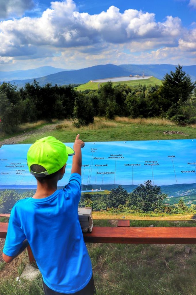 Dziecko w Beskidzie Małym - szczyt Kiczera, tablica prezentująca szczyty widoczne z Kiczery, widok na zbiornik retencyjny na Górze Żar, letnie popołudnie
