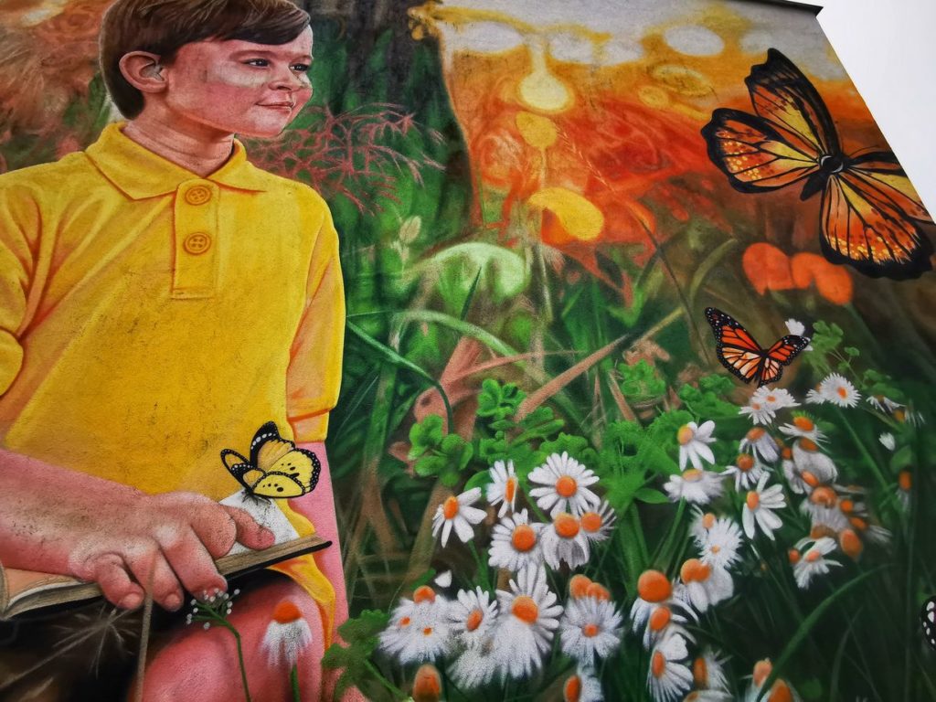 Mural w Jaworzu Średnim przedstawiający uśmiechniętego chłopca z książką na łące (kwiaty, motyle)