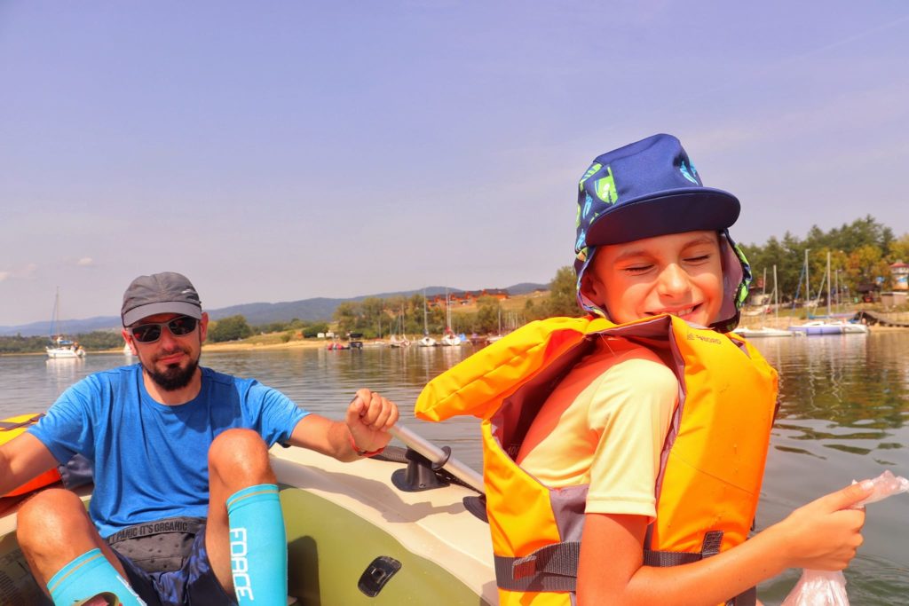 Szczęśliwe uśmiechnięte dziecko siedzące na dmuchanym pontonie z tatą - Jezioro Czorsztyńskie, w tle żaglówki, motorówki