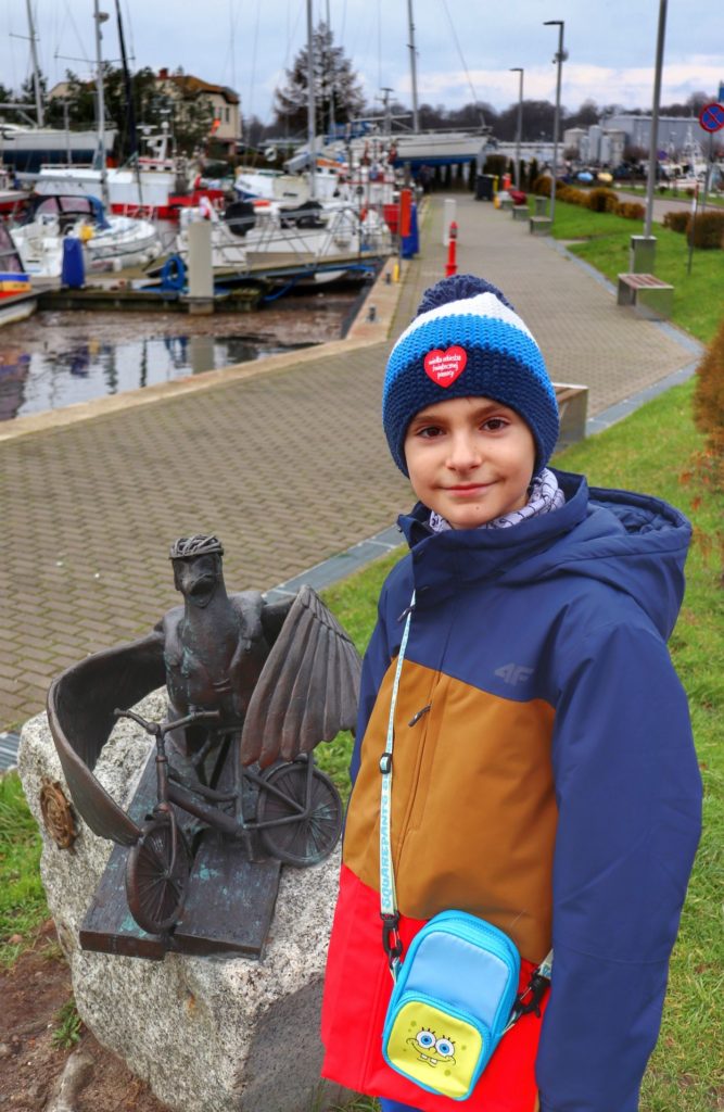 Zadowolone dziecko stojące przy rzeźbie mewy - Marian wsiadający na rower w Kołobrzegu - Port Jachtowy w Kołobrzegu