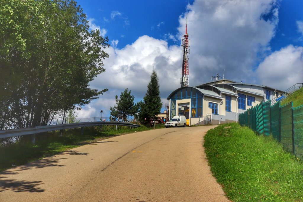 Budynek górnej stacji kolejki linowej na Górze Żar, szeroka asfaltowa droga na szlaku czerwonym, niebieskie niebo z białymi chmurami