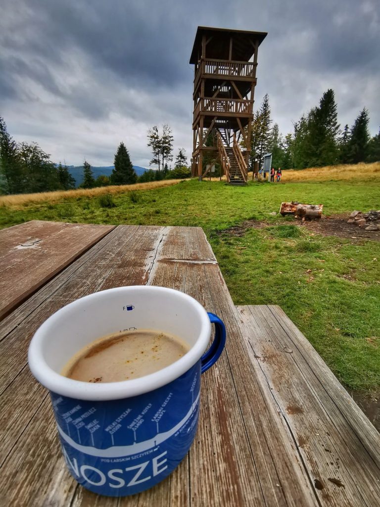 Drewniany blat, kubek z białą kawą, w tle wieża widokowa na Starym Groniu w Beskidzie Śląskim, ciemne niebo