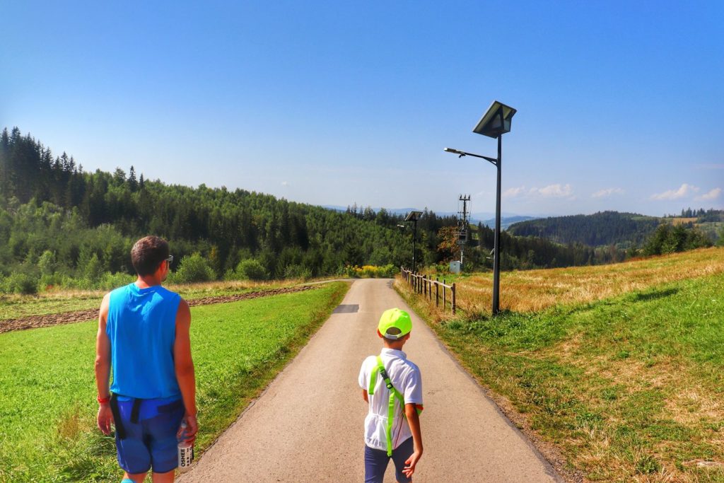 Turysta z dzieckiem idący asfaltową drogą - szlak żółty - Jaworzynka - Trójstyk granic - niebieskie niebo