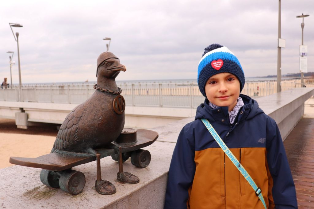 Rzeźba - Marian na deskorolce Kołobrzeg - rzeźba mewy, uśmiechnięte dziecko, w tle molo w Kołobrzegu oraz morze