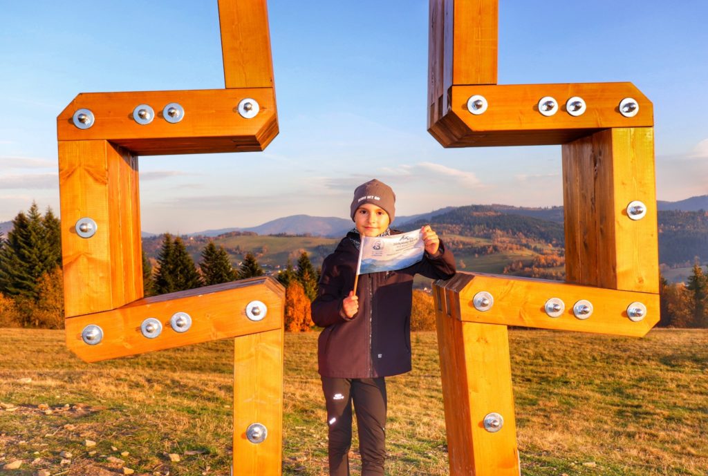 Szczyt Gruň, drewniany krzyż, uśmiechnięte dziecko z flagą akcji zdobywamy szczyty dla hospicjum