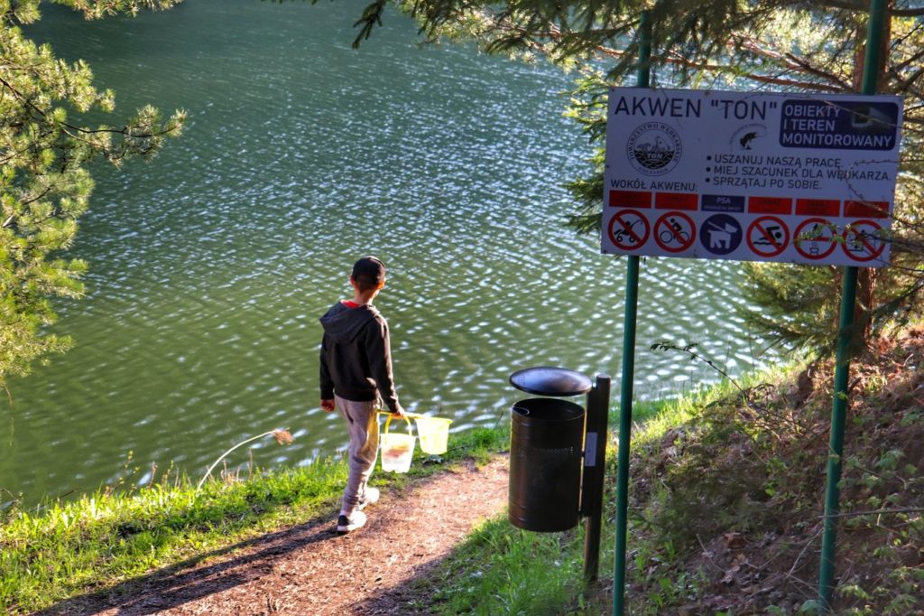Biała tabliczka informacyjna przy Jeziorze Ton w Goleszowie, dziecko, słoneczne popołudnie, dziecko idące wzdłuż brzegu jeziora