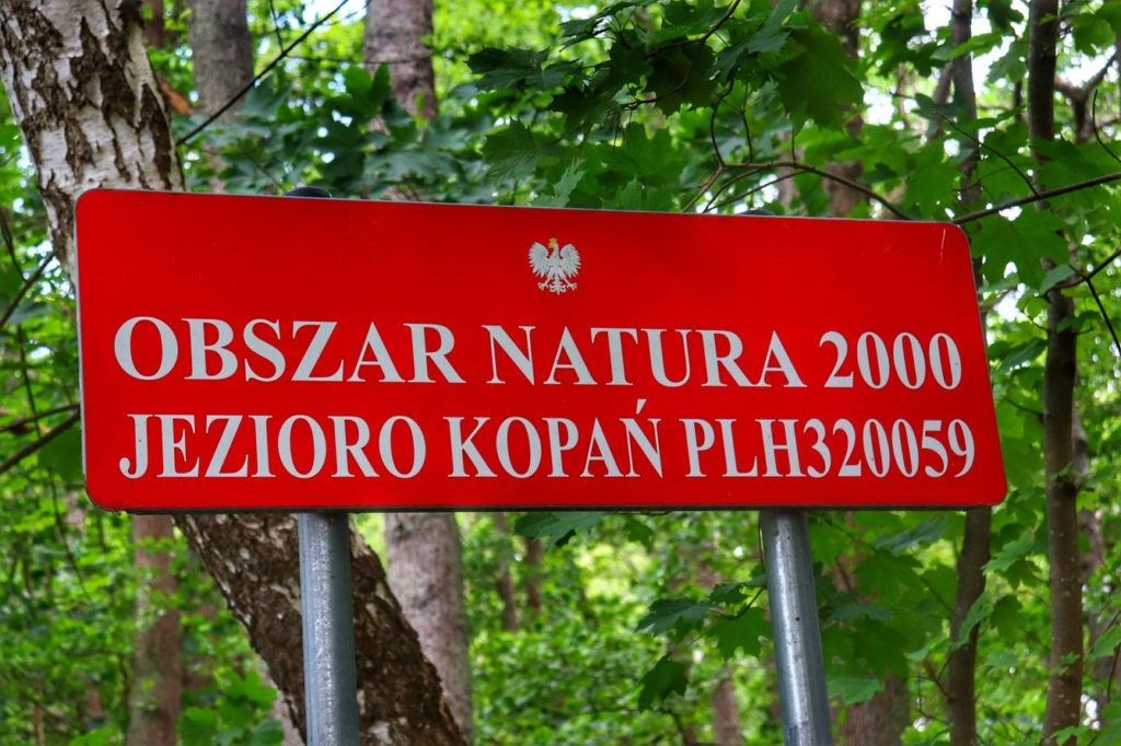 Czerwona tabliczka z białymi napisami - OBSZAR NATURA 2000 JEZIORO KOPAŃ - Wieś Wicie