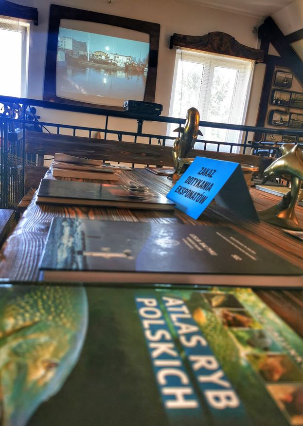 Stół, książki, atlasy ryb, film wyświetlany w Galerii Rybackiej w Jezierzanach