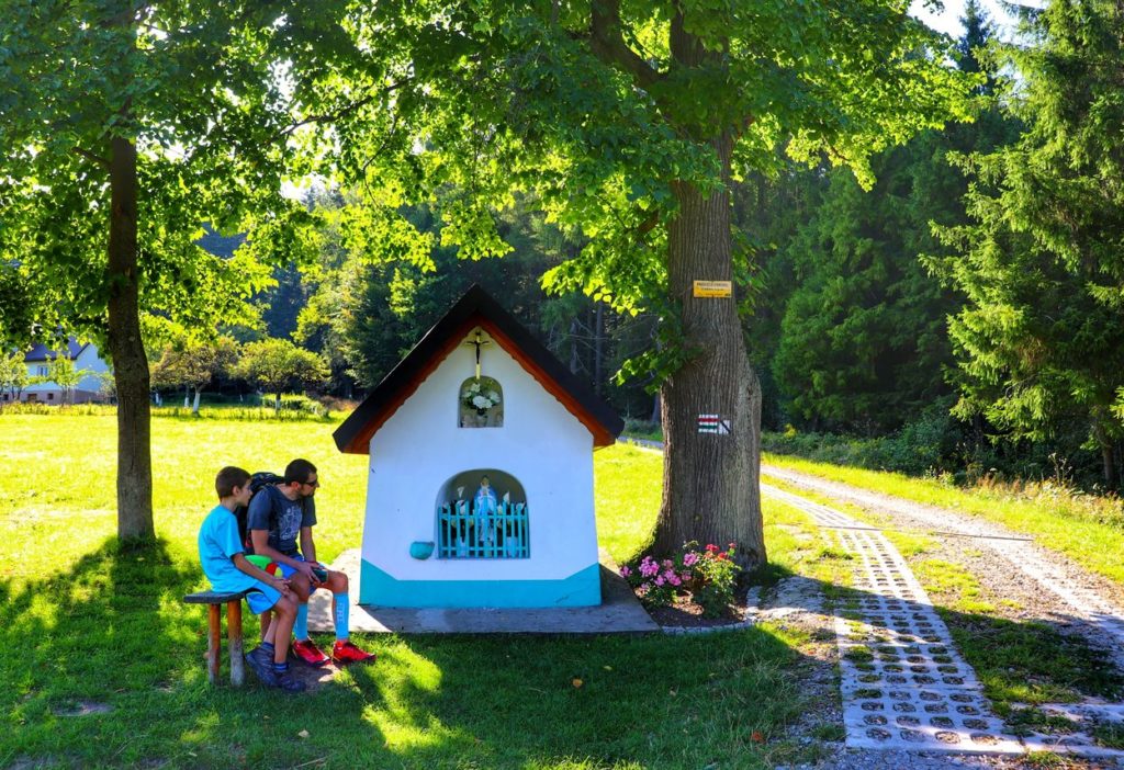 Przełęcz Carchel w Beskidzie Małym, murowana kapliczka Matki Boskiej, turysta z dzieckiem odpoczywający na drewnianej ławeczce przy murowanej kapliczce