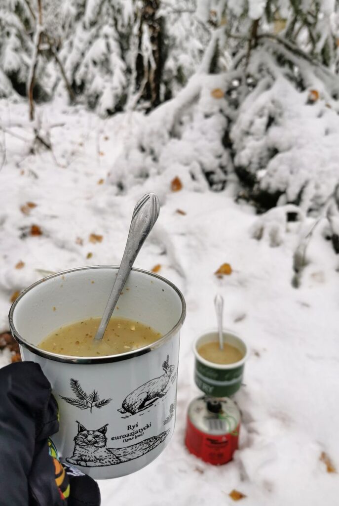 KUBEK EMALIOWANY z zupą, butla gazowa z palnikiem, na której położony jest kubek z żurkiem, beskidzki las, śnieg