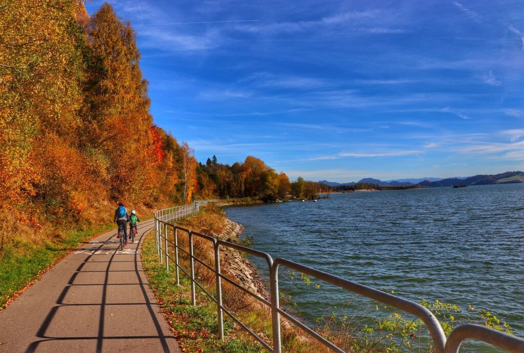 Rowerzyści jadący wzdłuż Jeziora Czorsztyńskiego, jesienna sceneria, niebieskie niebo, szlak rowerowy Velo Czorsztyn