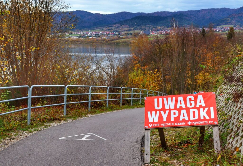Czerwona tablica z białym napisem UWAGA WYPADKI na rowerowej trasie Velo Czorsztyn - Falsztyn, widok na Jezioro Czorsztyńskie
