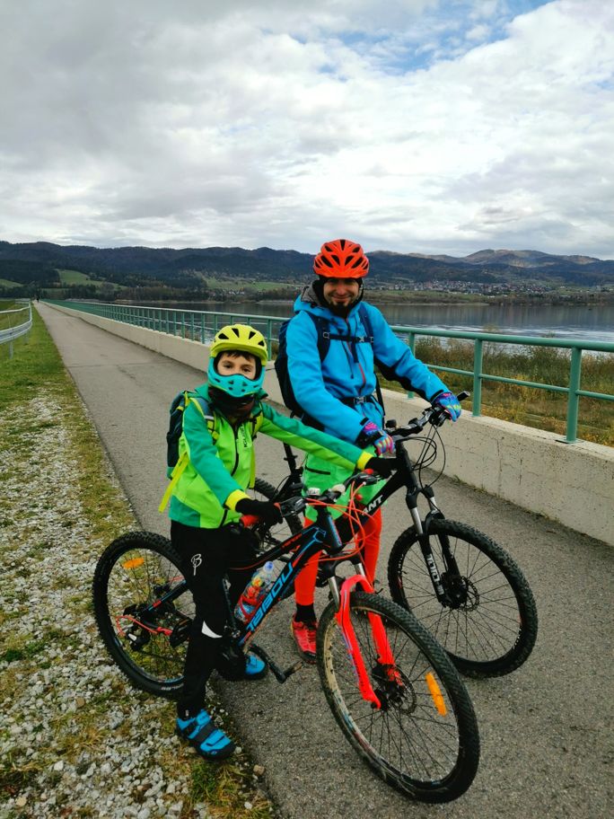 Uśmiechnięci rowerzyści, trasa Velo Czorsztyn biegnąca przy jeziorze, zachmurzone niebo
