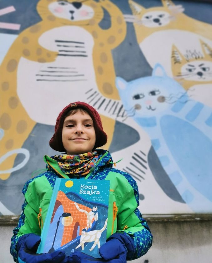 Uśmiechnięte dziecko na tle muralu Kocia Szajka, dziecko trzyma książkę pod tytułem "Kocia Szajka i napad na moście", miasto Cieszyn