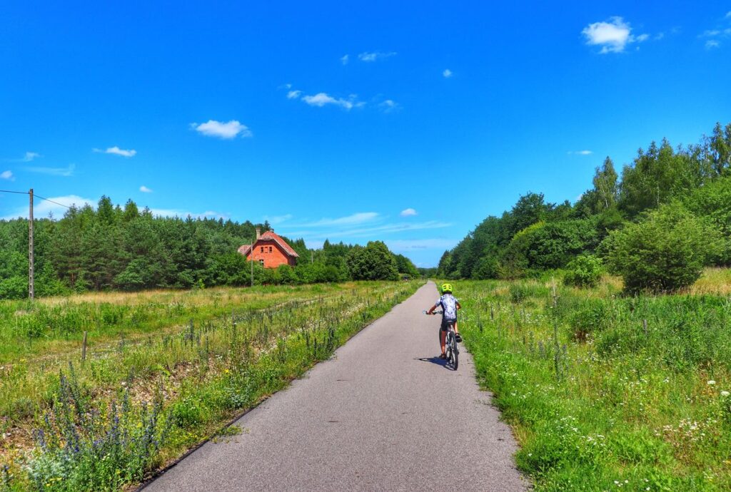 Dziecko na rowerze na trasie Rowerem po Kolei na Mazurach, na horyzoncie stacja kolejowa OCHÓDNO