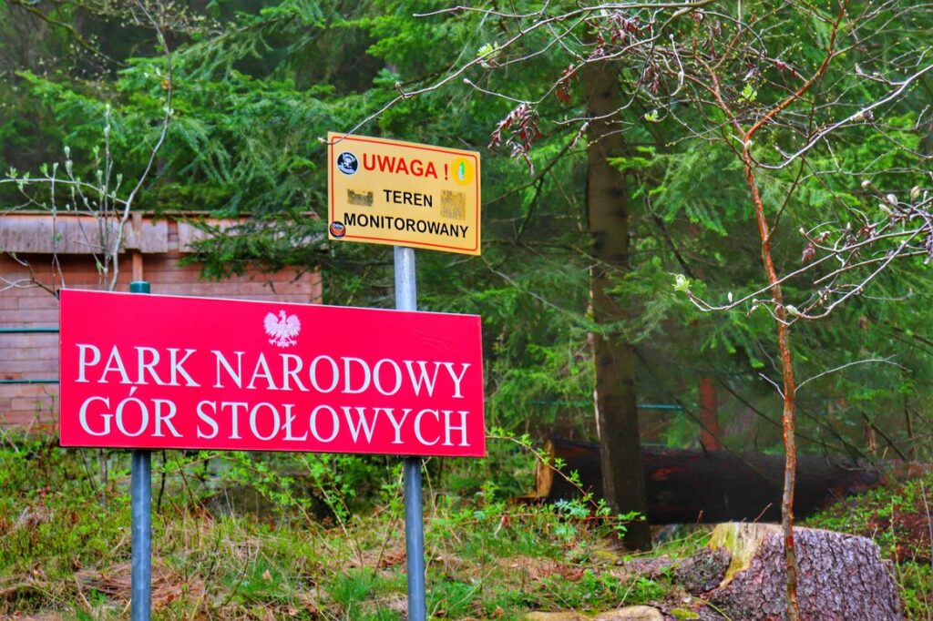 Czerwona tablica z białym napisem - PARK NARODOWY GÓR STOŁOWYCH, żółta tabliczka z czerwonym napisem - UWAGA! i czarnym napisem - TEREN MONITOROWANY, trasa na Szczeliniec