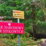 Czerwona tablica z białym napisem - PARK NARODOWY GÓR STOŁOWYCH, żółta tabliczka z czerwonym napisem - UWAGA! i czarnym napisem - TEREN MONITOROWANY, trasa na Szczeliniec