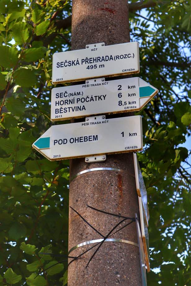 Biała tablica wisząca na drzewie z napisem Sečská přehrada (rozc.) oraz drogowskazami zielonego szlaku