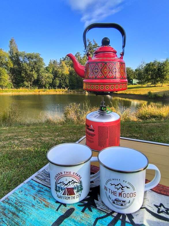 Dwa białe emaliowane kubki z motywem biwaku i podróży, turystyczny stół z butlą gazową oraz czerwonym czajniczkiem w etniczny wzór, a w tle przepiękny staw Hetlik w Skuteč