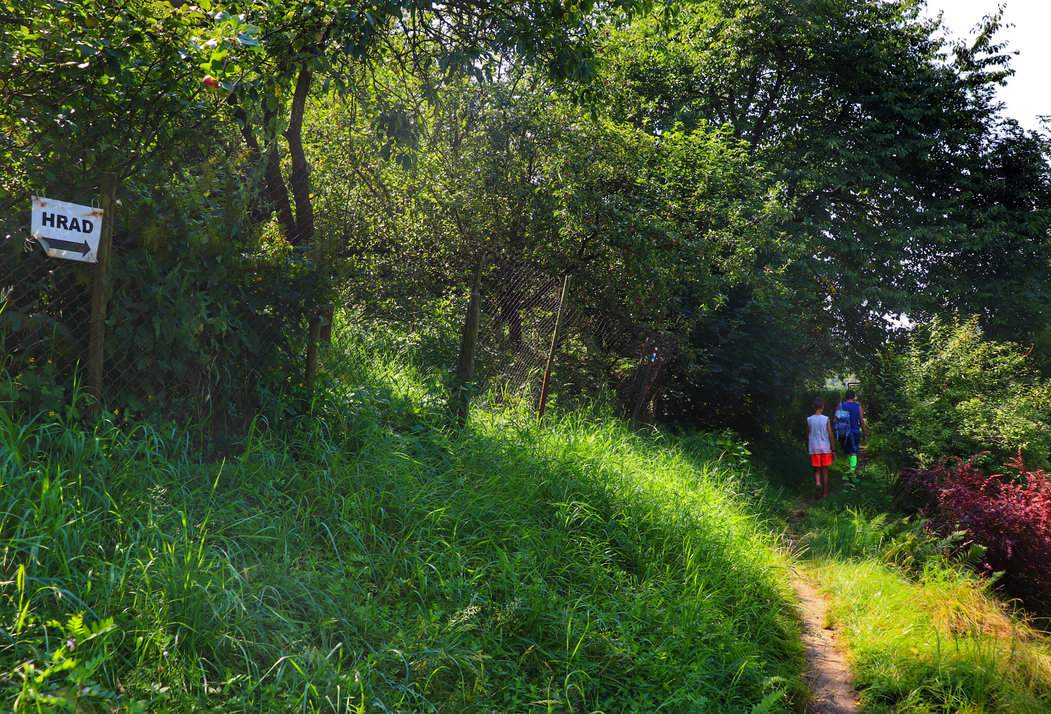 Biała tabliczka wisząca na płocie wskazująca kierunek do ruin zamku Brníčko w Czechach; wąska ścieżka i turyści - dziecko z tatą