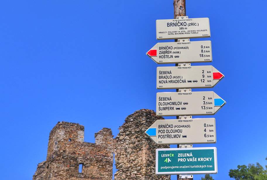 Słup z drogowskazami przy ruinach zamku Brníčko, biała tabliczka z napisem „Brníčko - ruiny”, w tle ruiny zamku