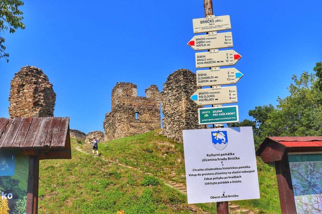 Słup z drogowskazami przy ruinach zamku Brníčko, biała tabliczka z napisem „Brníčko - ruiny”, w tle turysta wchodzący po schodach oraz ruiny zamku