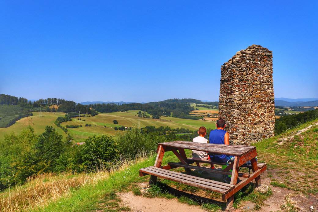 Turysta z dzieckiem przy drewnianym stole z ławkami, z widokiem rozpościerającym się z ruin zamku Brníčko w Czechach, podczas wspaniałego, letniego dnia