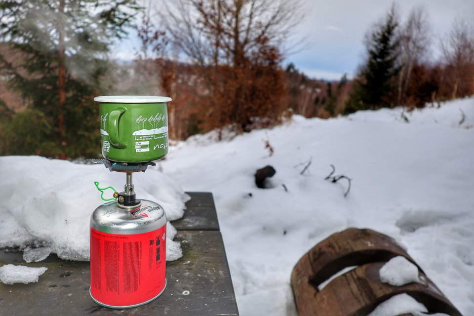Emaliowany kubek z gotującą się wodą na palniku gazowym, położony na zaśnieżonym drewnianym stole - przerwa na kawę na słowackim szlaku