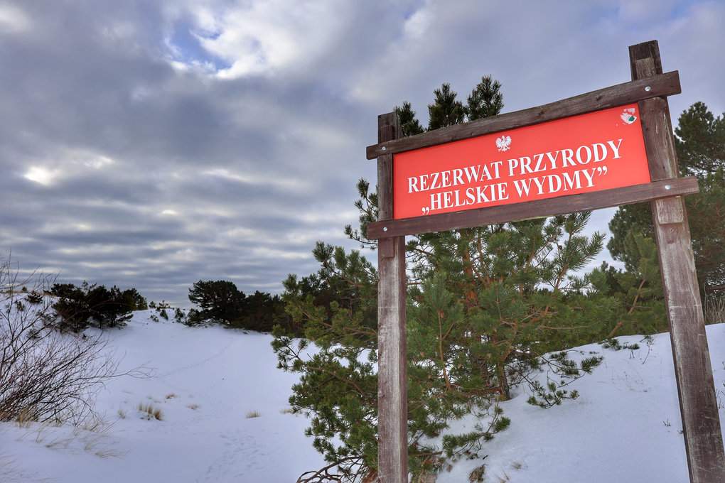 Czerwona tabliczka z białym napisem: „Rezerwat przyrody Helskie Wydmy”