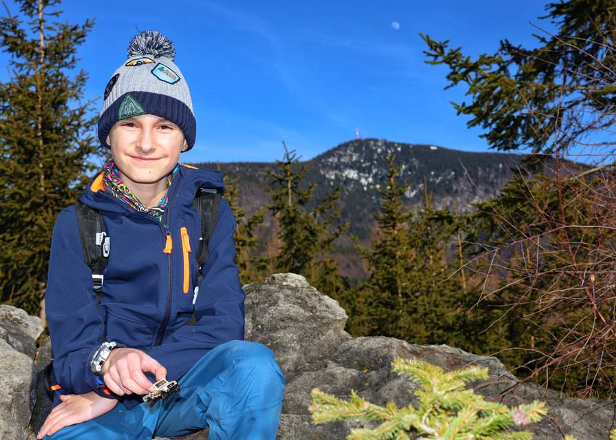 Uśmiechnięte dziecko przy formacji skalnej Liščí díry w Beskidzie Morawsko-Śląskim, w tle najwyższy szczyt pasma - Łysa Góra (Łysa Hora) - i niebieskie niebo